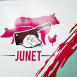 Logo de la Boucherie Junet : silhouettes d’un bœuf, d’un cochon et d’une poule en rouge, avec une hachoir de boucher stylisé posée sur une tranche de viande, et le nom “JUNET” en lettres capitales rouges en dessous.